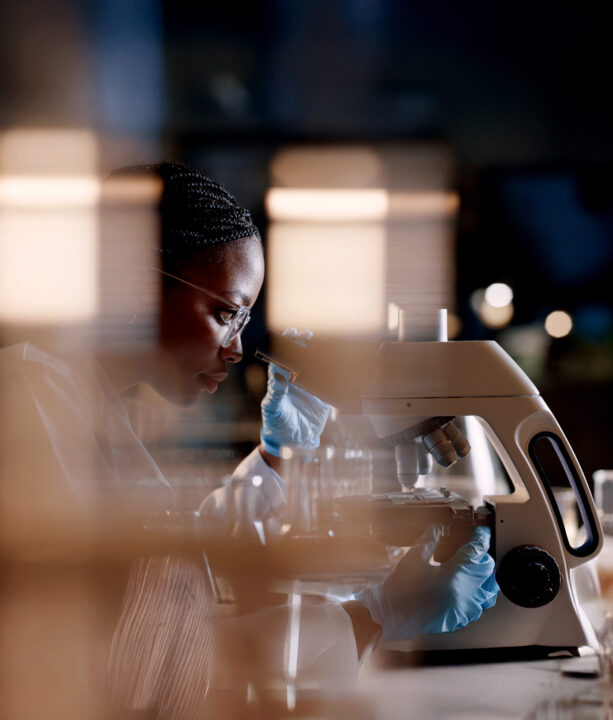 A BSc (Hons) Bioscience with Digital Technologies student wearing safety glasses and blue gloves examines a sample under a microscope in a laboratory, viewed through blurred glass, creating a focused, professional atmosphere.