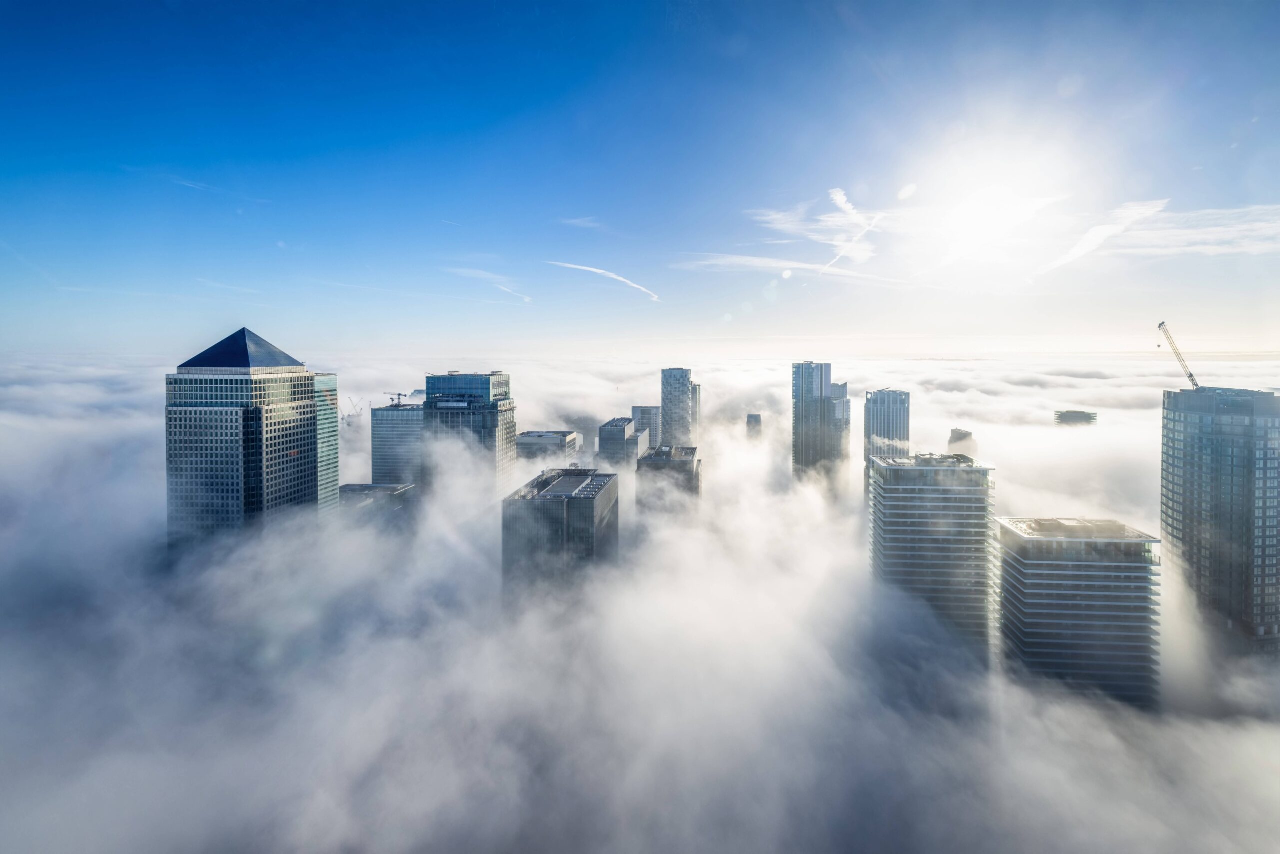 Aerial view of skyscrapers emerging through thick fog under a bright blue sky, with sunlight shining and soft clouds surrounding the buildings—an inspiring scene for anyone pursuing a Business degree London.