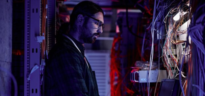 A person with long hair and glasses works on a laptop in a server room, surrounded by tangled cables and electronic equipment, illuminated by blue and red lights—immersed in their Data Science BSc studies.