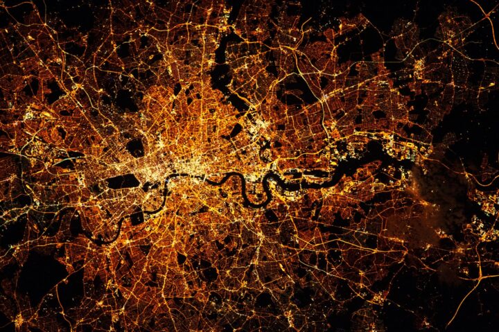 Aerial night view of a brightly lit city with a winding river at its center, streets glowing orange and yellow—like the interconnected pathways one studies in an Economics BSc (Hons), revealing a vibrant network against the dark background.