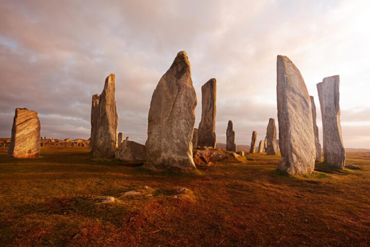 Large, weathered standing stones form a circle on grassy, sunlit ground under a cloudy sky, creating an ancient and mysterious atmosphere reminiscent of sites studied in BA Hons History London courses.