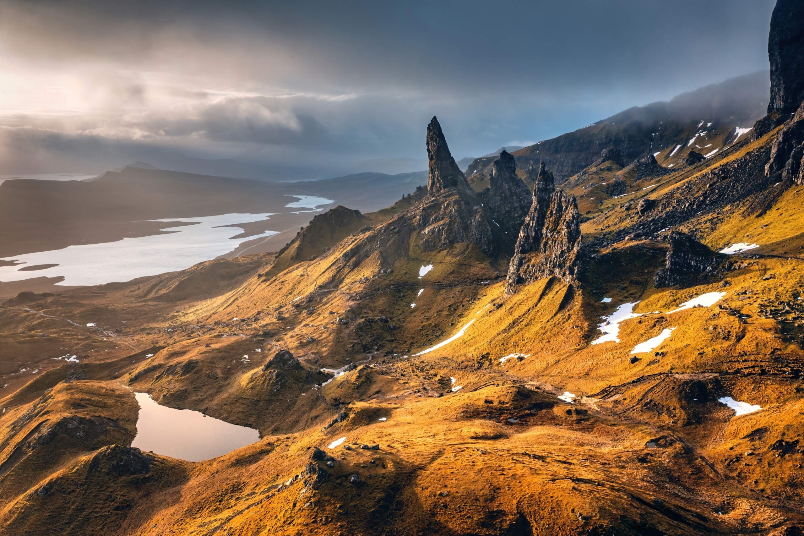 Dramatic rocky pinnacles rise above golden grassy hills with patches of snow, overlooking a winding lake under a cloudy sky in this rugged, mountainous landscape—ideal inspiration for anyone studying BA Hons Philosophy London.