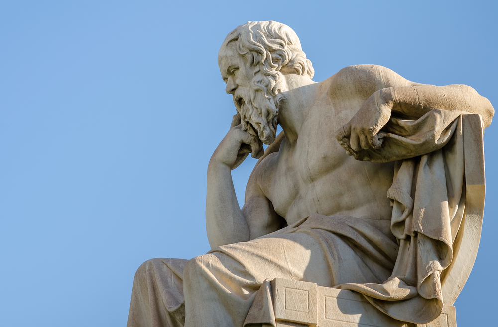 A marble statue of a seated philosopher, depicted in deep thought. The figure has a long beard and is partially draped with fabric. The statue is set against a clear blue sky.
