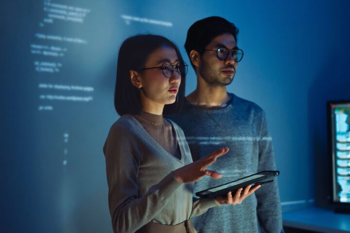 Two people wearing glasses stand in a dimly lit room with computer code projected on the wall behind them; one holds a tablet and gestures as they discuss their BSc (Hons) Computer Science & Business London project, both focused intently off camera.