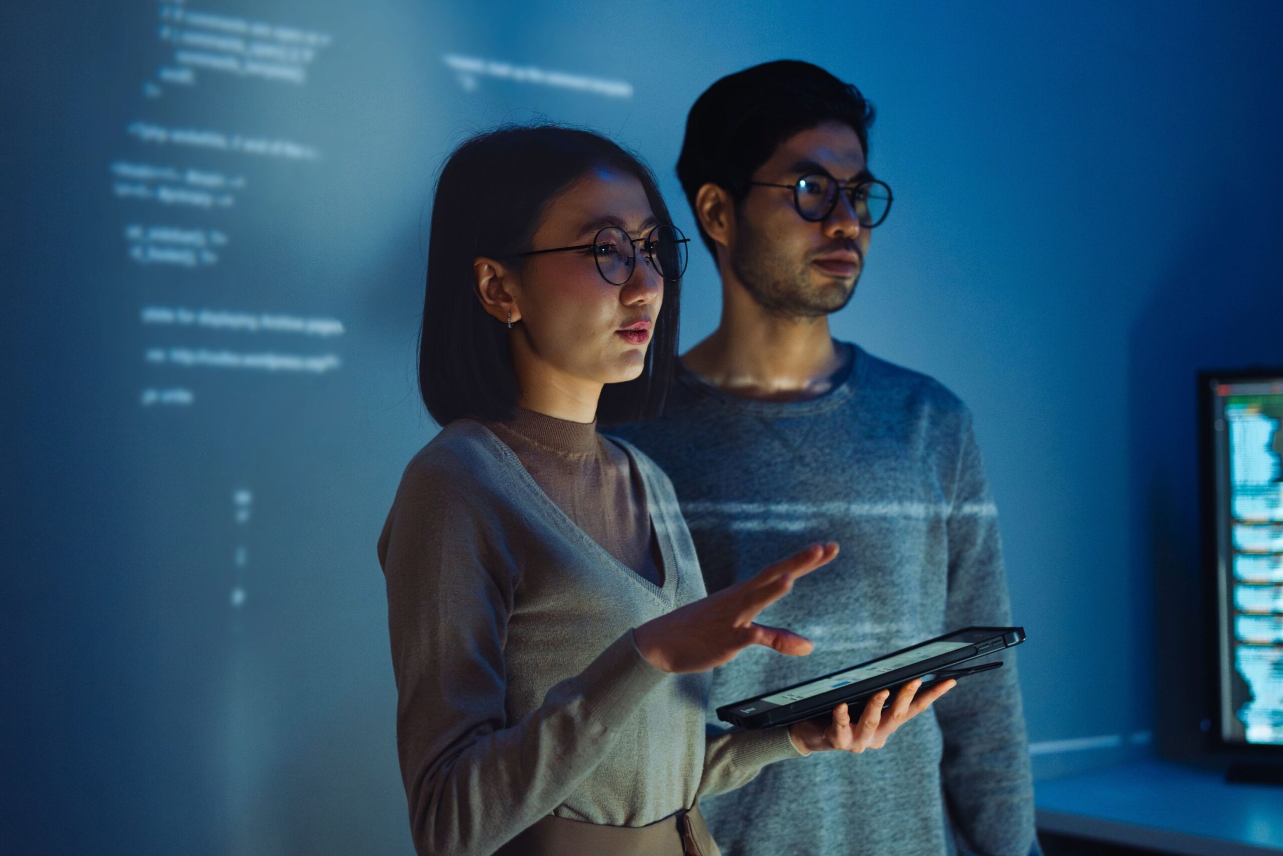Two people wearing glasses stand in a dimly lit room with computer code projected on the wall behind them; one holds a tablet and gestures as they discuss their BSc (Hons) Computer Science & Business London project, both focused intently off camera.
