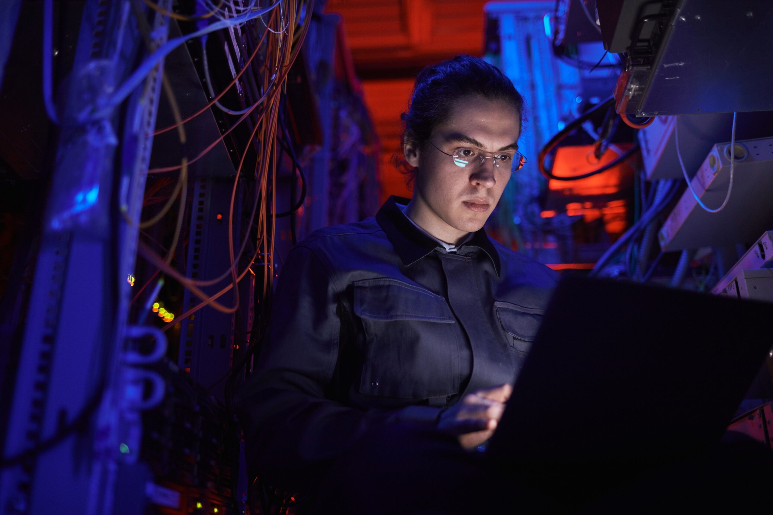 A person wearing glasses and a dark jacket works on a laptop, surrounded by network cables and server equipment in a dimly lit room with blue and red lighting, immersed in the intersection of politics & data science.