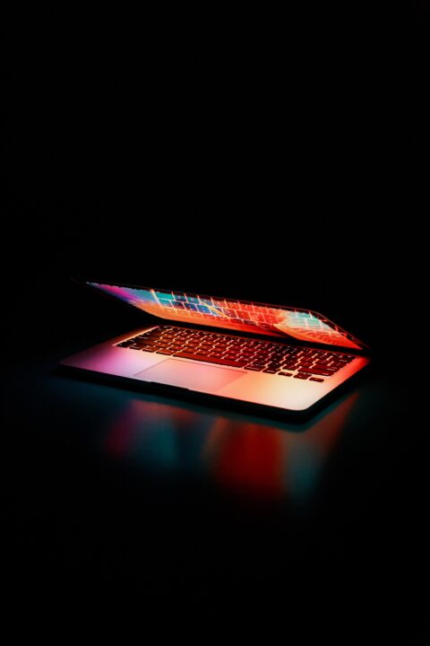 A partially opened laptop glows in the dark, displaying a colorful gradient of reds, pinks, and blues on the screen and keyboard. The light reflects softly on the surface beneath it, creating a vivid, atmospheric scene.