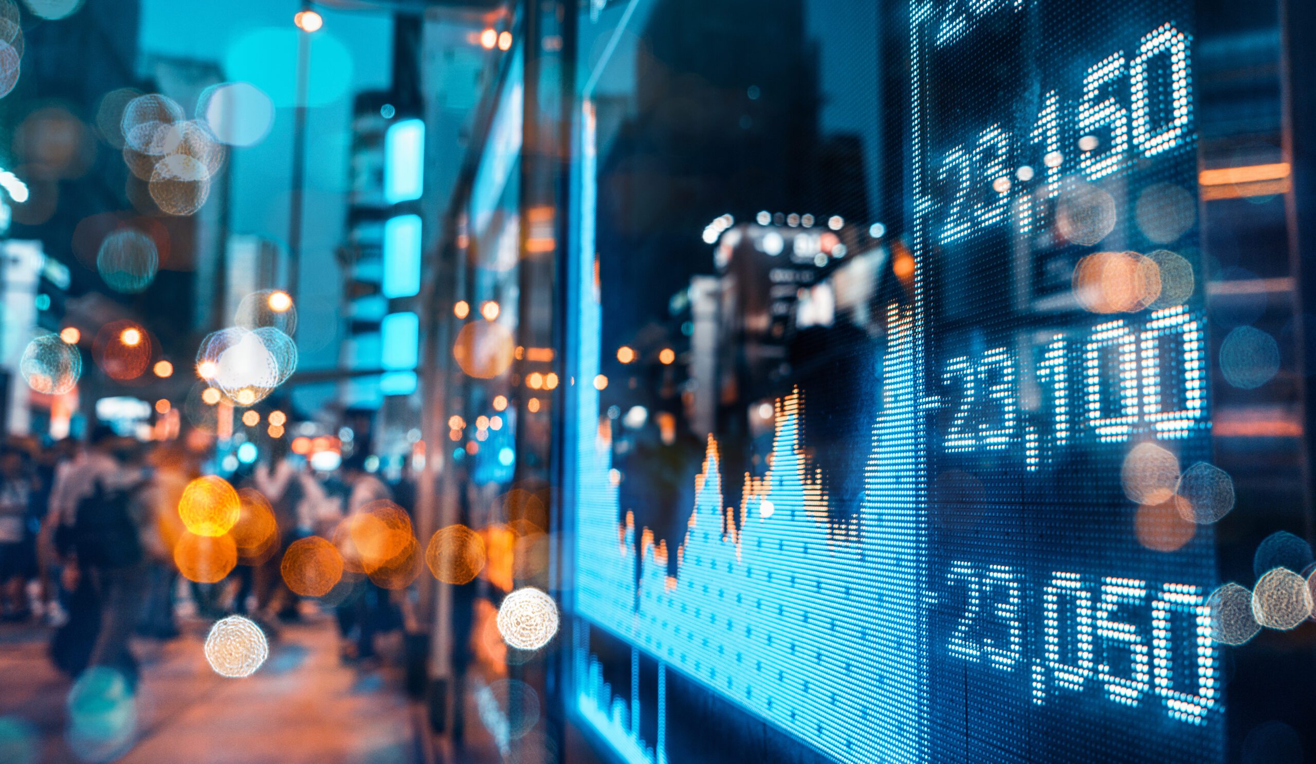 A digital display on a city street shows fluctuating financial graphs and numbers, with a blurred background of pedestrians and illuminated buildings at night.