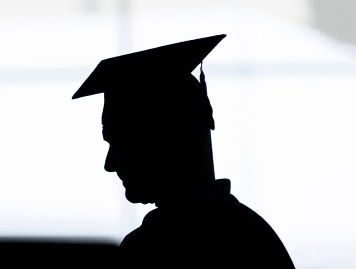 Silhouette of a person wearing a graduation cap, shown in profile against a bright background.