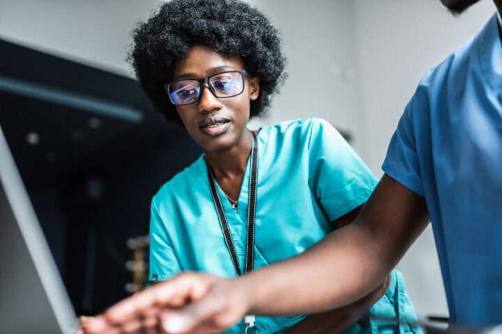 A Clinical Trials Specialist in teal scrubs and glasses looks at a computer screen while another person in medical attire points at the monitor during a discussion in a clinical setting.