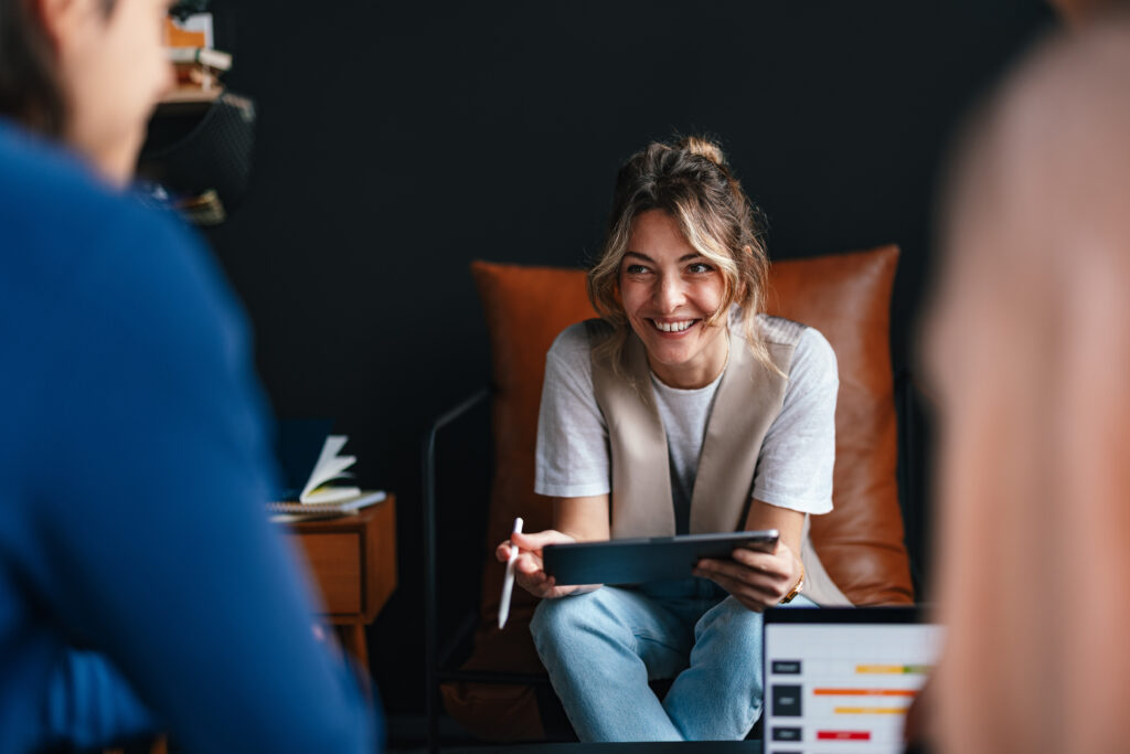 An Accounting Finance Manager sits on a brown chair, smiling as she holds a tablet and stylus, engaging with two people in a meeting. A laptop with a chart appears in the foreground of the modern office setting.