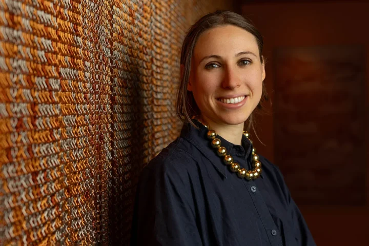 Calypso Newman is smiling, wearing a black shirt and a chunky gold necklace. She stands in front of a textured wall with a warm, amber tone. The lighting casts soft shadows, creating a cozy ambiance.