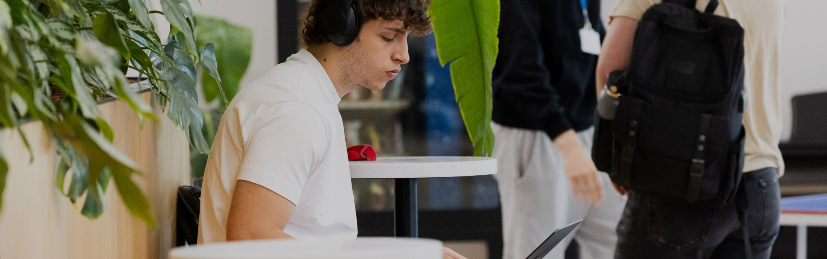 A person wearing headphones is sitting at a small table, using a laptop. Another person with a backpack stands nearby. Green leaves are visible in the foreground, adding a touch of nature to the indoor scene.