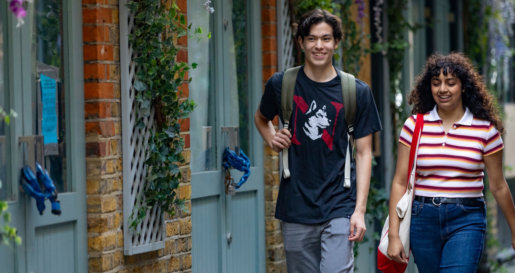 A young man and woman walking side by side in a leafy courtyard. The man wears a black t-shirt with a graphic and carries a backpack. The woman wears a striped shirt and jeans. They appear to be smiling and enjoying the day.