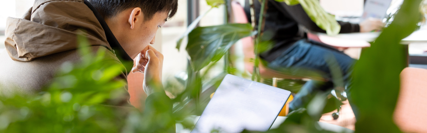 A person sitting among green plants, focused on looking at a laptop screen. They are wearing a brown jacket and have their hand near their mouth. Another person is seated in the background, facing a computer.