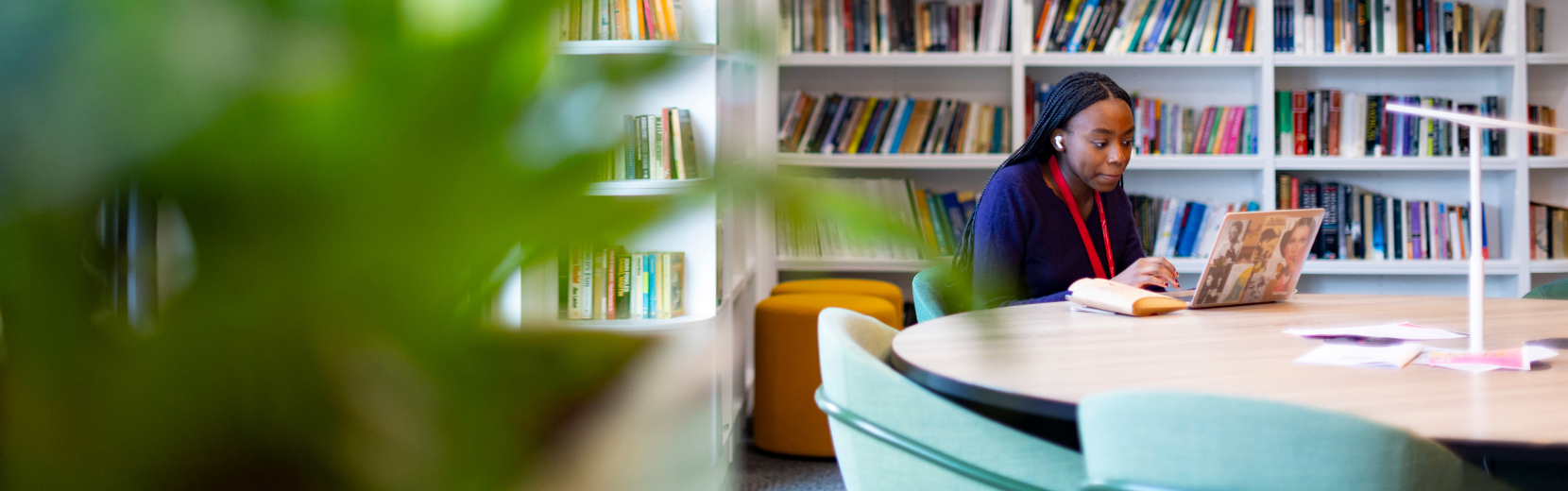 A person sits at a round table in a library, working on a laptop. Bookshelves filled with books line the wall in the background. A green plant is visible in the foreground, slightly blurred.
