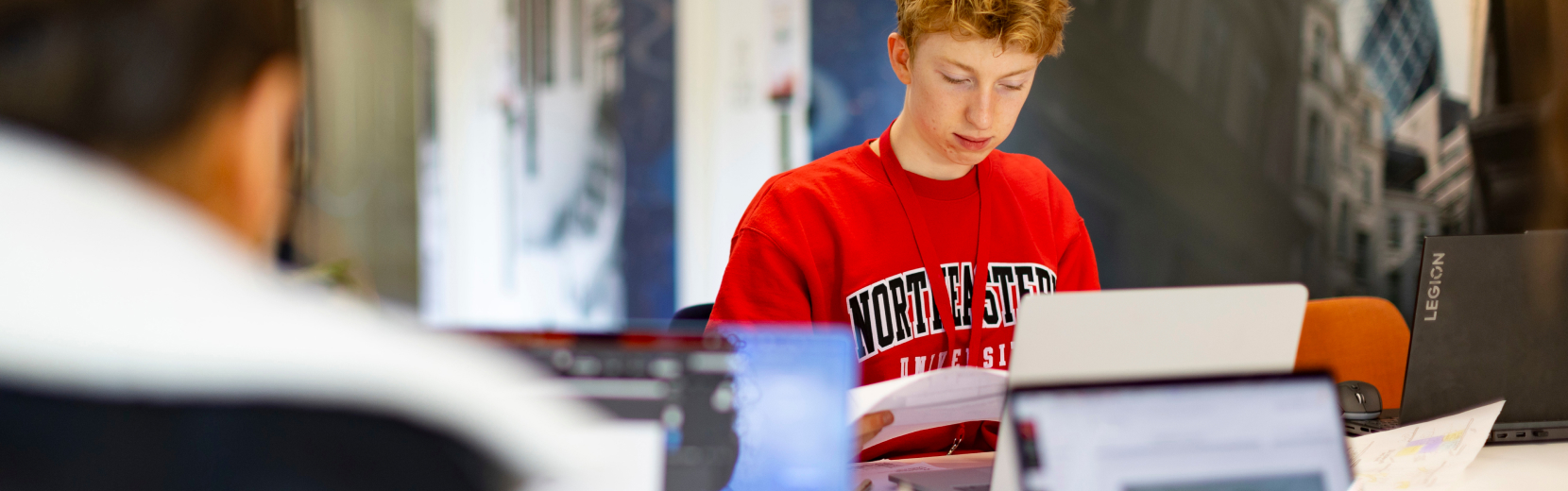 A person in a red "Northwestern" sweatshirt is focused on reading a paper while using a laptop in a room with other computers. The background shows blurred figures and office decor.