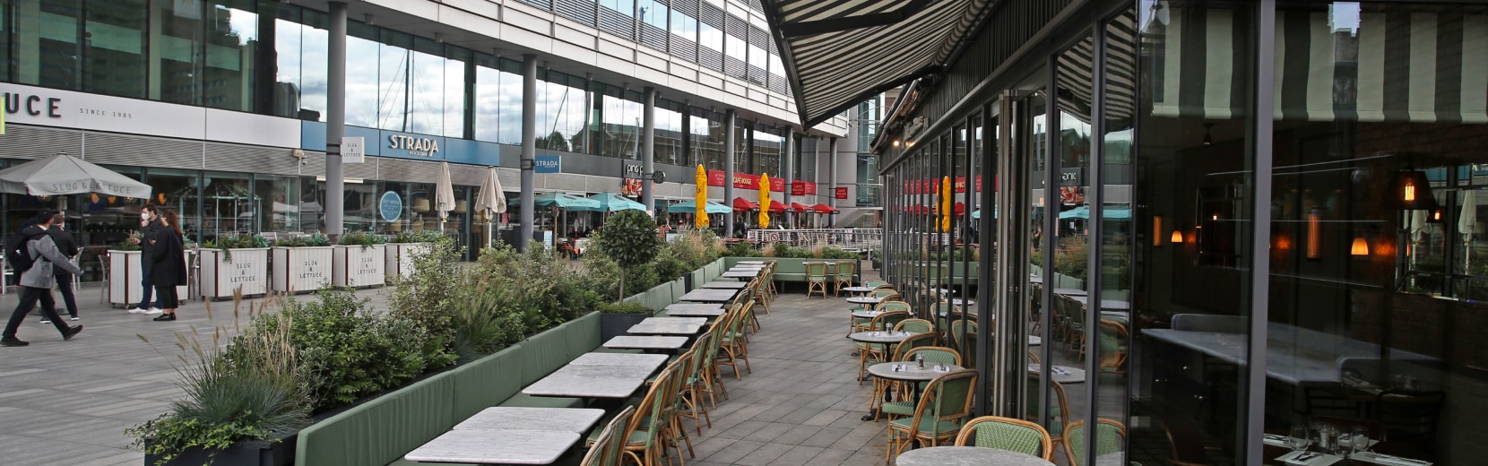 Outdoor cafe with empty tables and chairs along a pedestrian street, surrounded by greenery. People are walking nearby, and storefronts are visible in the background under a cloudy sky.