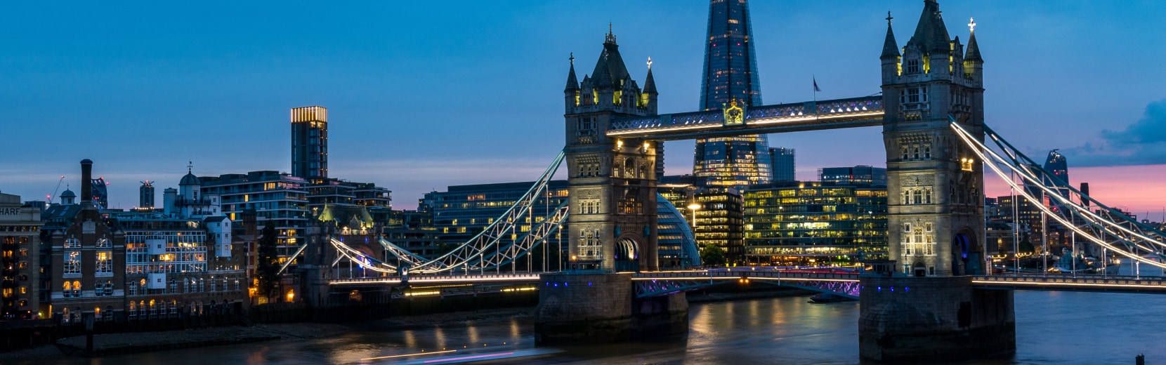 A panoramic view of Tower Bridge lit up at dusk in London, with the Shard skyscraper in the background. The city skyline is reflected in the River Thames, and buildings are illuminated against the evening sky.