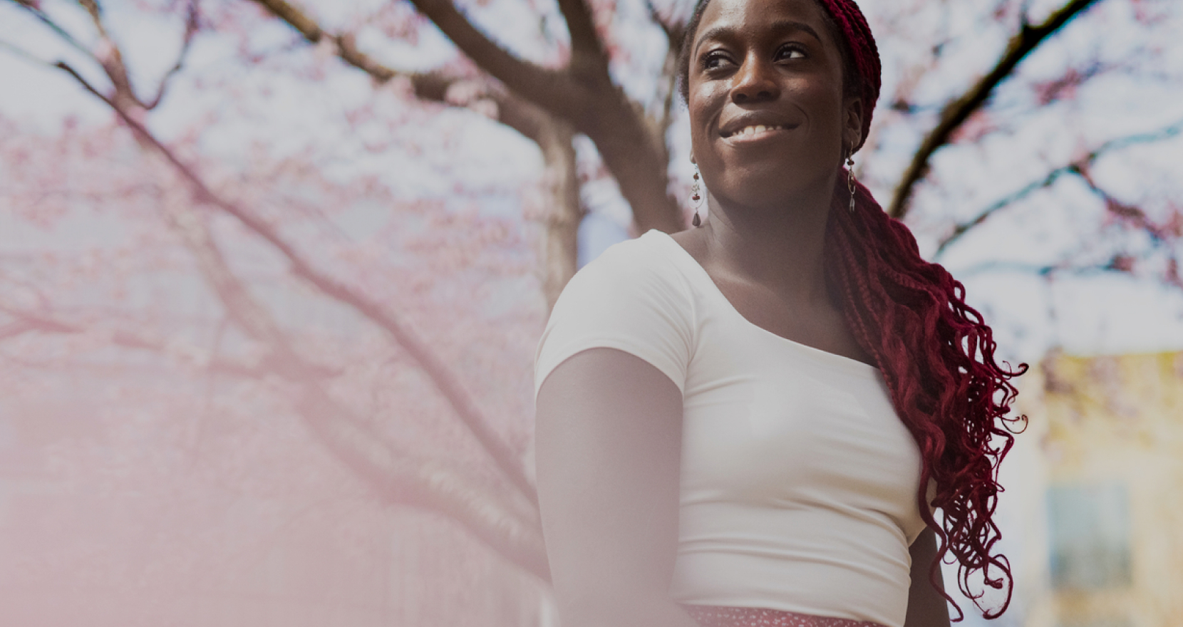 A woman with red braided hair smiles while standing outdoors under pink cherry blossom trees. She is wearing a white top and patterned skirt. The background features blooming branches and a soft, blurred effect.