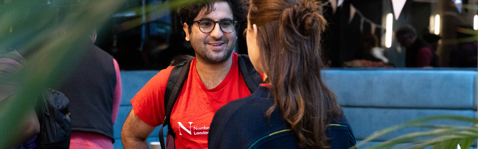 A man in a red shirt is smiling and talking to a woman with brown hair tied back. The background is blurred, showing a casual indoor setting with plants and dim lighting.