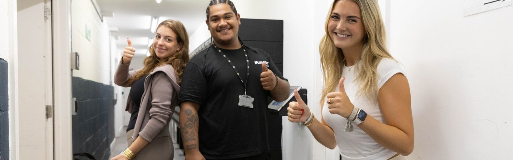 Three people stand in a hallway giving thumbs up and smiling. The person on the left wears a brown sweater, the person in the middle wears a black shirt and lanyard, and the person on the right wears a white shirt. The hallway has white walls with a dark stripe.