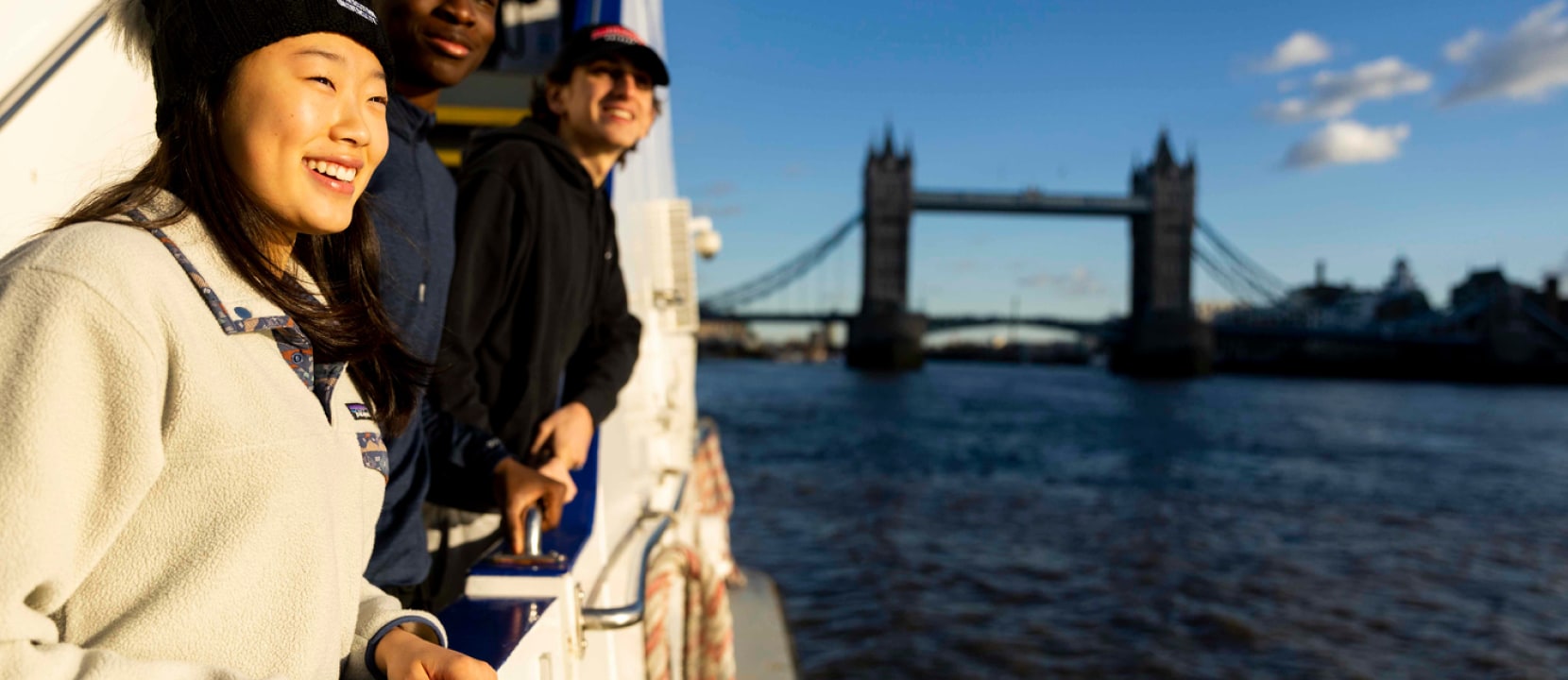 Three people enjoy a sunny day on a riverboat with a view of Tower Bridge in the background. They are smiling and looking out over the water, suggesting a sense of adventure and joy.