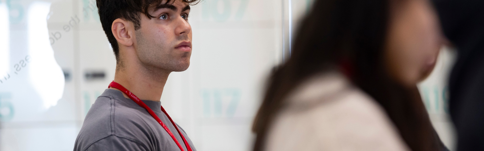 A man with curly hair wearing a gray shirt and a red lanyard stands in a room, looking attentively to the side. The background is blurred, showing another person and a wall with soft lighting.