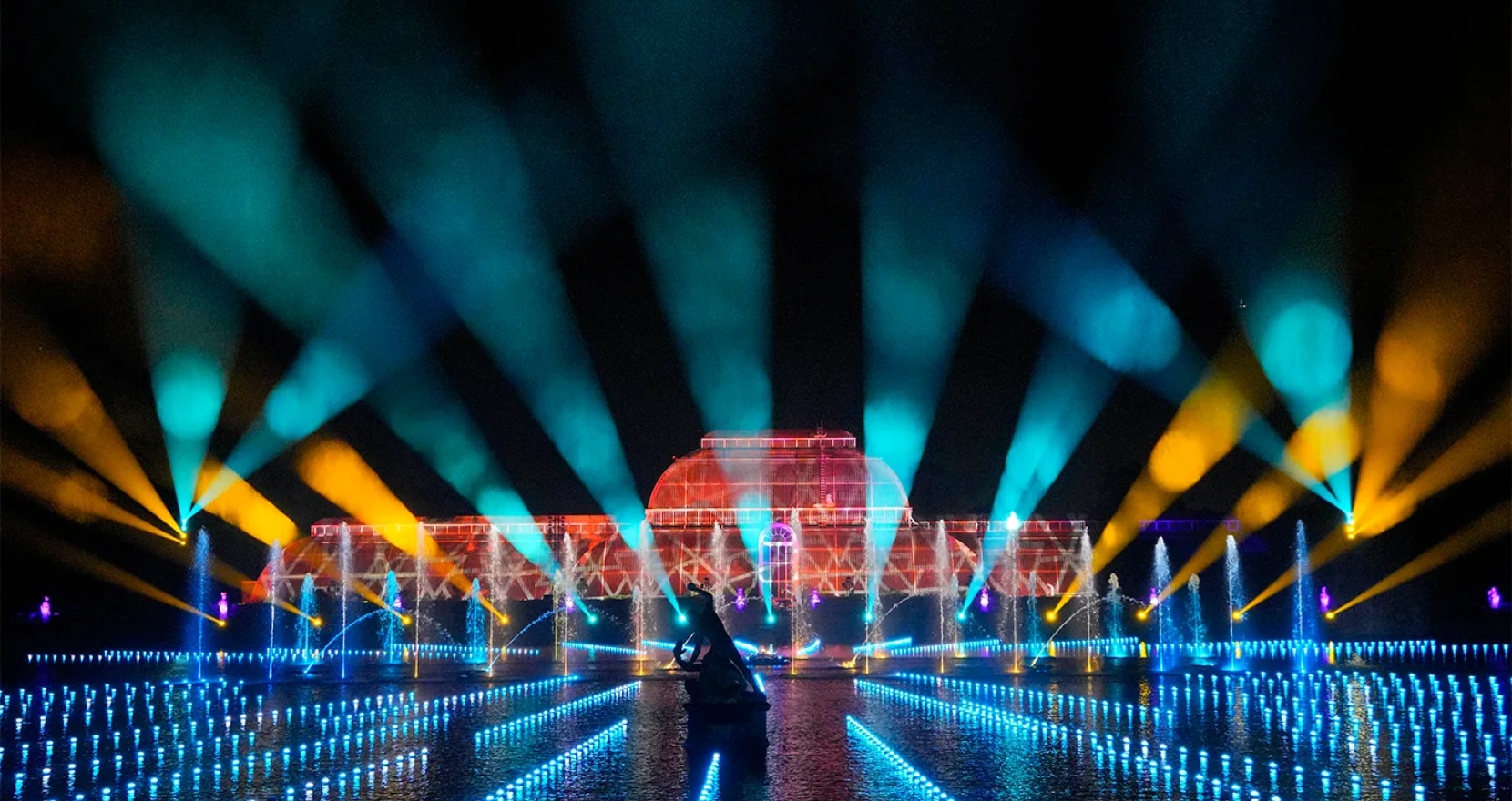 A vibrant light show over water features colorful beams and reflections, illuminating a large, symmetrical structure. Blue, orange, and green lights crisscross the scene, creating a dynamic and festive atmosphere.