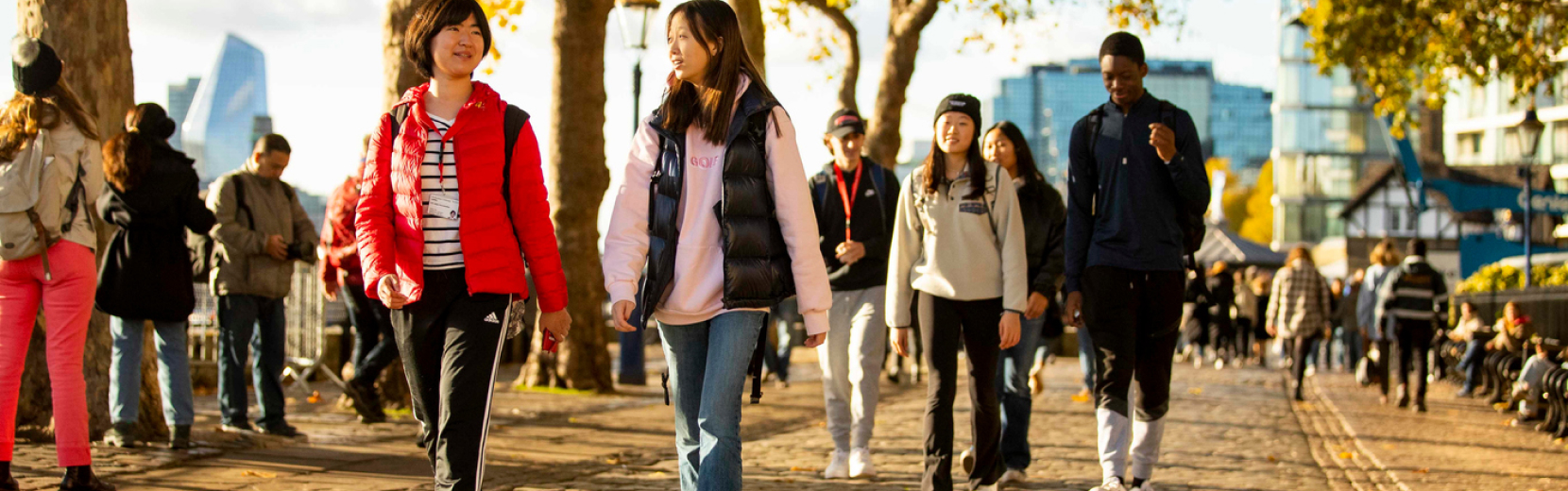 A group of young people walk along a tree-lined path on a sunny day. They are dressed casually, chatting and smiling. In the background, a crowd of people and urban buildings are visible.