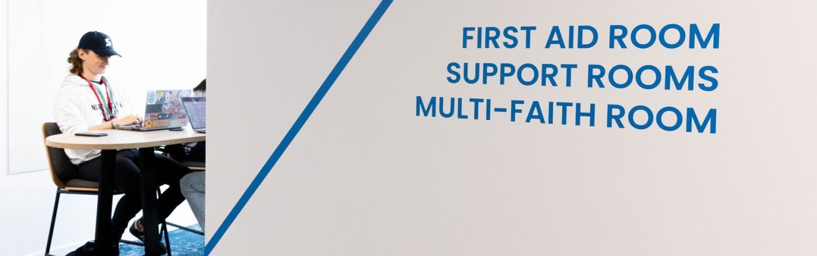 A person sits working on a laptop in a room with a sign on the wall listing facilities: "First Aid Room, Support Rooms, Multi-Faith Room.