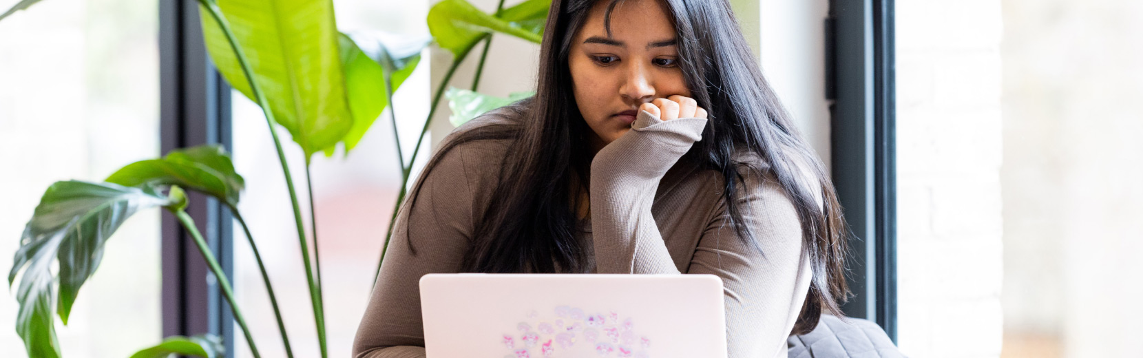 A woman with long dark hair wearing a brown sweater sits at a table, looking at her laptop with a thoughtful expression. She's surrounded by large green plants and natural light coming through expansive windows.