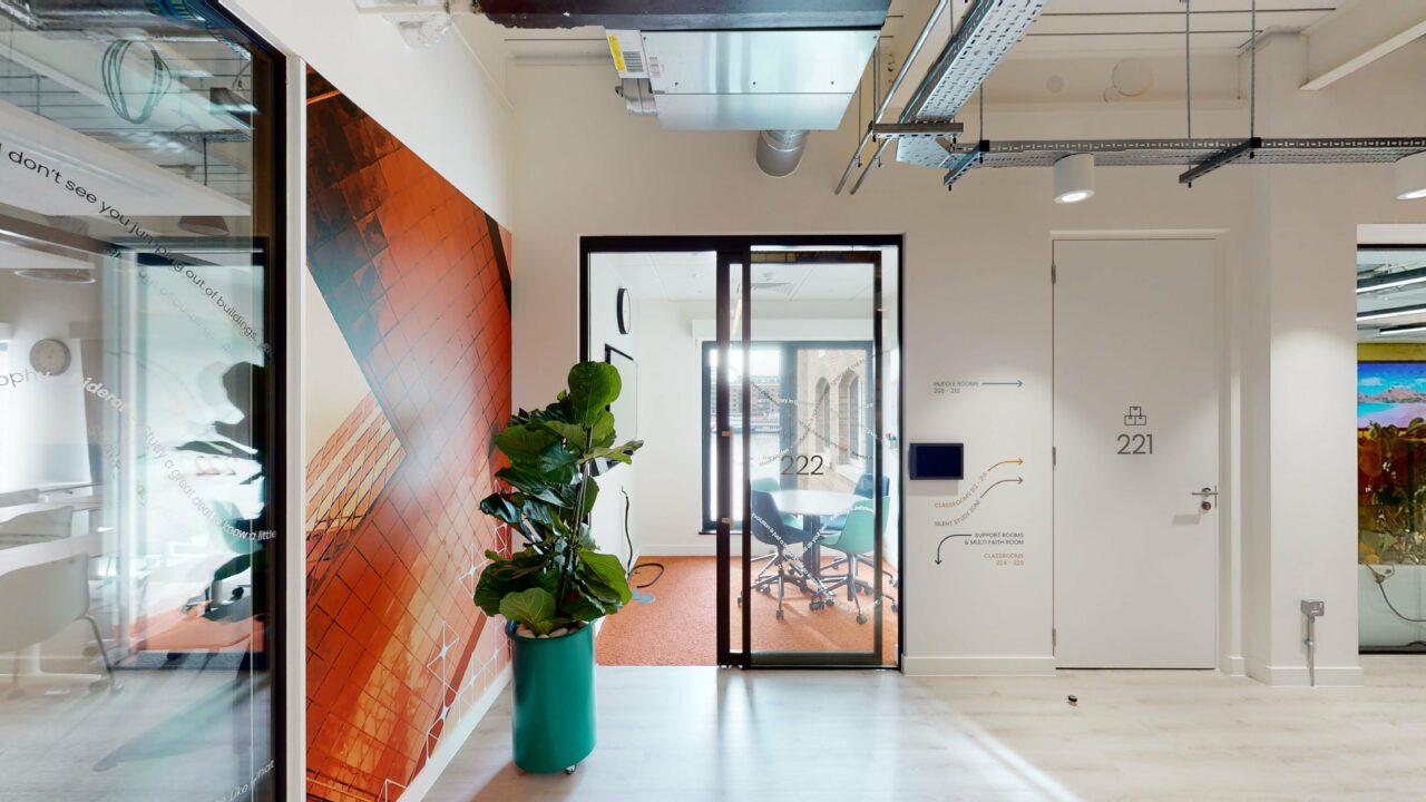 Modern office hallway with glass doors leading to a meeting room, labeled 221. The room features a table with chairs. A large plant is near a colorful wall mural. Visible ceiling ducts and light wooden flooring emphasize the contemporary design.