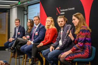 A panel of six people sitting and talking during an event at Northeastern University London. A woman on the right holds a microphone. A presentation screen is partially visible in the background.
