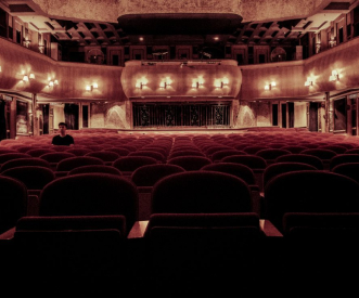 A dimly lit theater with red velvet seats, mostly empty, except for a single person sitting alone in the middle. The stage is illuminated with warm lights, enhancing the vintage interior and ornate balcony design.