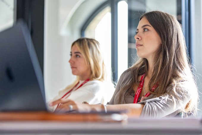 Two women seated at a desk with laptops, focusing intently. The woman in the foreground wears a gray sweater and red lanyard, while the woman behind her has blonde hair. They appear to be in a modern office or classroom setting.