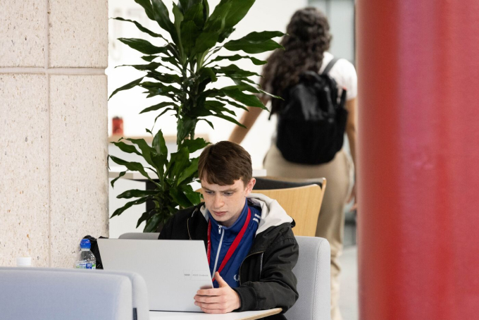 A young person with a laptop sits at a table in an indoor setting. They have a focused expression. Behind them, another person with a backpack walks by. A tall potted plant stands nearby, and a red pillar is partially visible in the foreground.