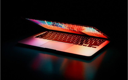 A partially open laptop emits vibrant colors from its screen against a dark background, reflecting hues of red, blue, and orange on the surface below.