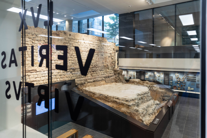A glass-walled room displays an ancient stone wall excavation. Artifacts are showcased on the surrounding walls. The word "VERITAS" is partially visible on the glass. Bright lighting and windows illuminate the exhibit from above.