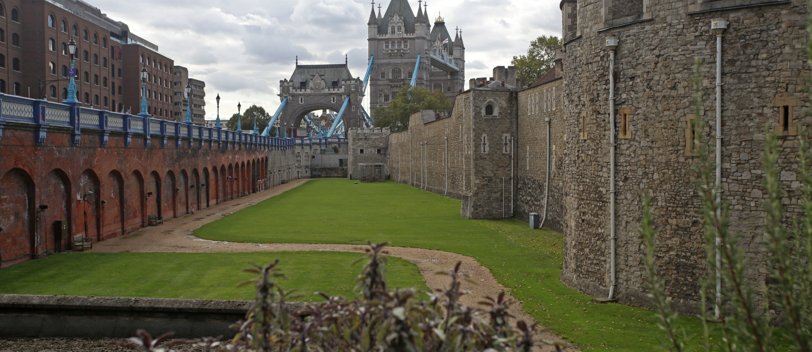 View of the Tower of London's outer walls and surrounding green area, with the iconic Tower Bridge in the background on a cloudy day. Buildings and structures showcase historical architecture.