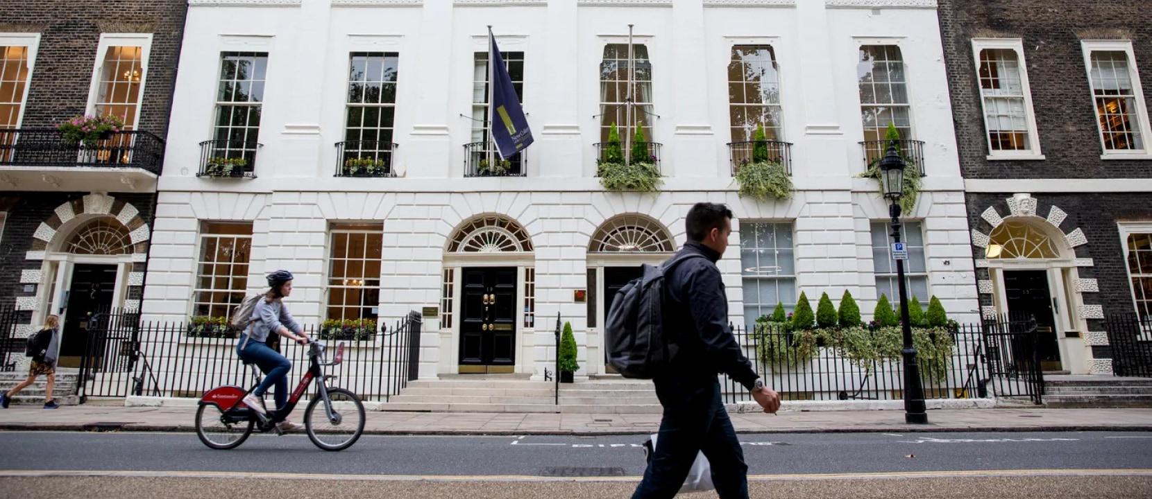 A person wearing a backpack walks past a large building with a white facade. A cyclist rides next to them. The building has arched windows with potted plants and a flag above the entrance. It's flanked by dark brick structures.