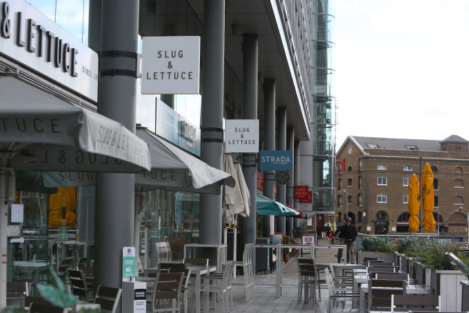 Outdoor seating area of the Slug & Lettuce restaurant with tables and chairs under umbrellas. The scene includes other restaurants and shops along the walkway, with a brick building and bright yellow outdoor umbrellas in the background.