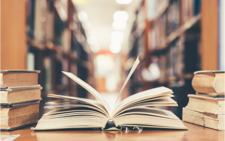 An open book sits on a wooden table between stacks of closed books in a library. Shelves filled with more books are visible in the background, creating a cozy and studious atmosphere.