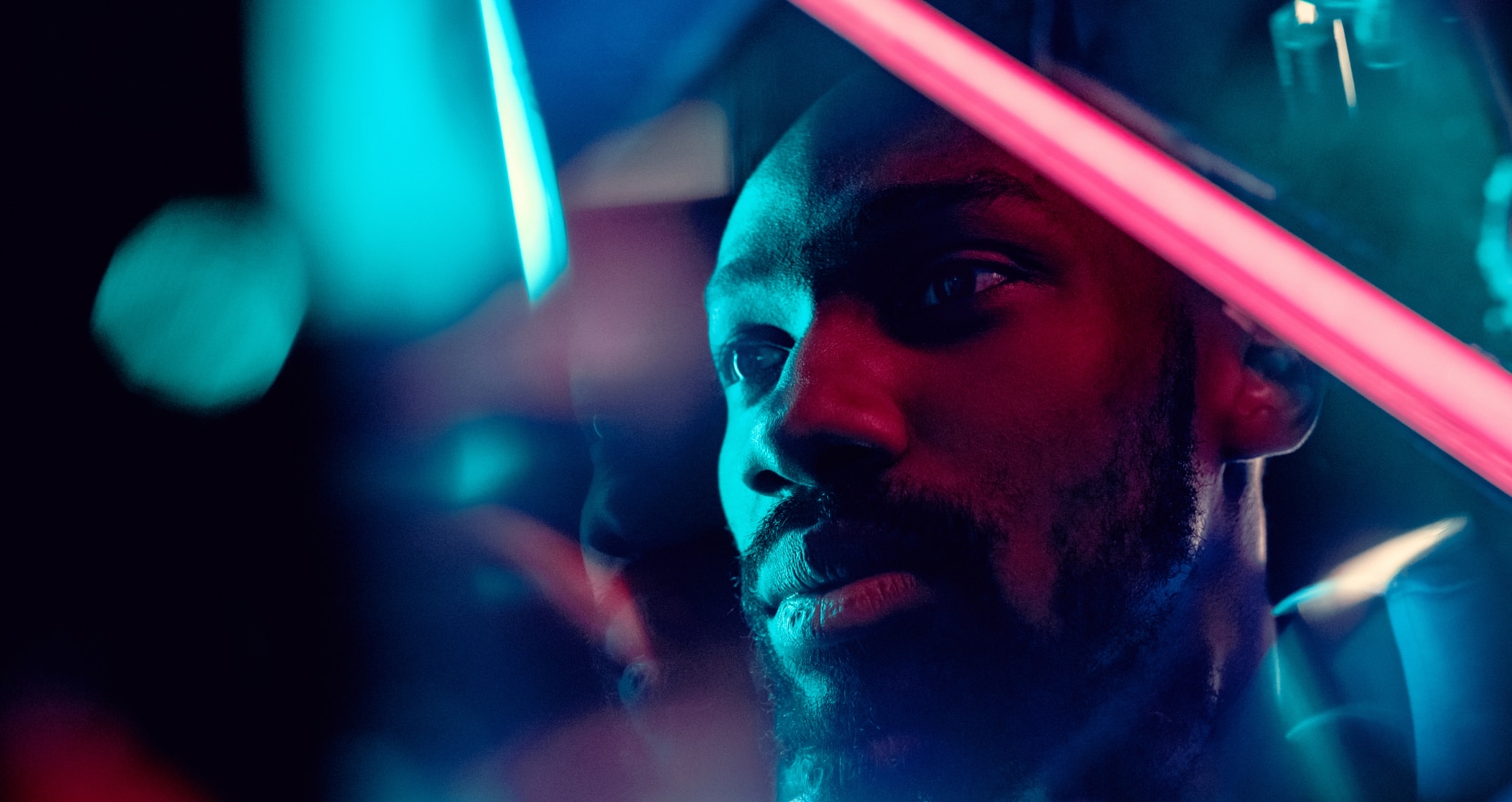 A person with a contemplative expression is illuminated by vibrant neon lights in shades of blue and pink. The image has a futuristic, abstract feel, with reflections and blurred elements creating a sense of depth and complexity.