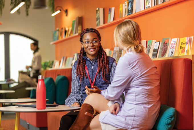 Two people sitting on a couch in a colorful library or lounge area, engaged in conversation. One person is holding a pen, and a red water bottle is on the table. Shelves with books line the orange wall behind them.