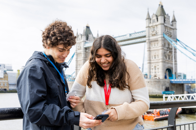 Two people stand by a riverfront railing, smiling and looking at a phone together. They wear lanyards and jackets. The Tower Bridge is visible in the background, indicating they are in London. The sky is cloudy.