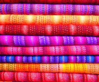 A stack of colorful woven textiles featuring bright stripes and gradient patterns in hues of red, orange, pink, purple, and yellow. The textures and vibrant colors create a warm, inviting appearance.