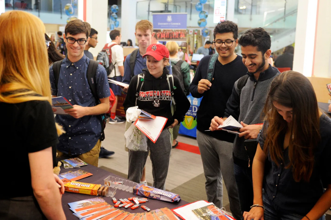 Students gather around a booth at a university event, interacting with a representative. They hold brochures and appear engaged. The background shows more students and informational displays.