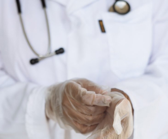 A healthcare professional wearing a white lab coat and a stethoscope prepares to put on transparent gloves. The focus is on the hands and the coat, capturing the readiness for medical procedures.
