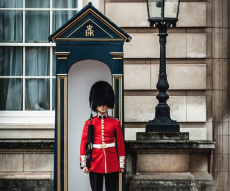 A Royal Guard in a red uniform and bearskin hat stands in a black and gold sentry box outside a historic building. A streetlamp is visible nearby.